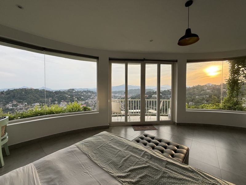 Guest bedroom with curved corner windows framing a sunset over the Kandy hills at The Summit Kandy