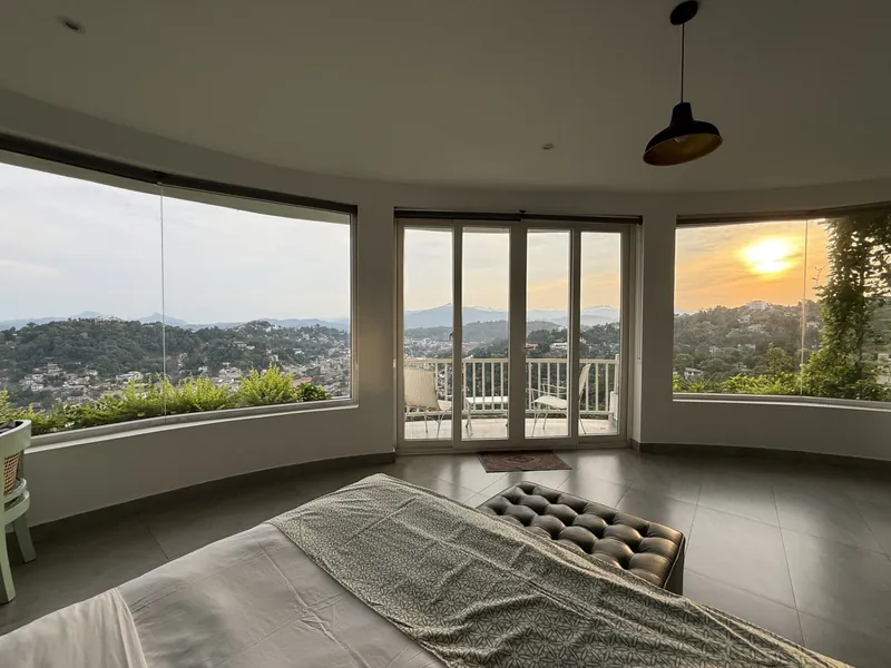 Guest bedroom with curved corner windows framing a sunset over the Kandy hills at The Summit Kandy